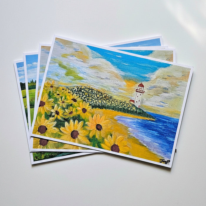 A stack of printed art cards or postcards lying on a light gray surface. The top card shows a painted coastal landscape with yellow sunflowers, blue water, a white lighthouse, and a cloudy sky.