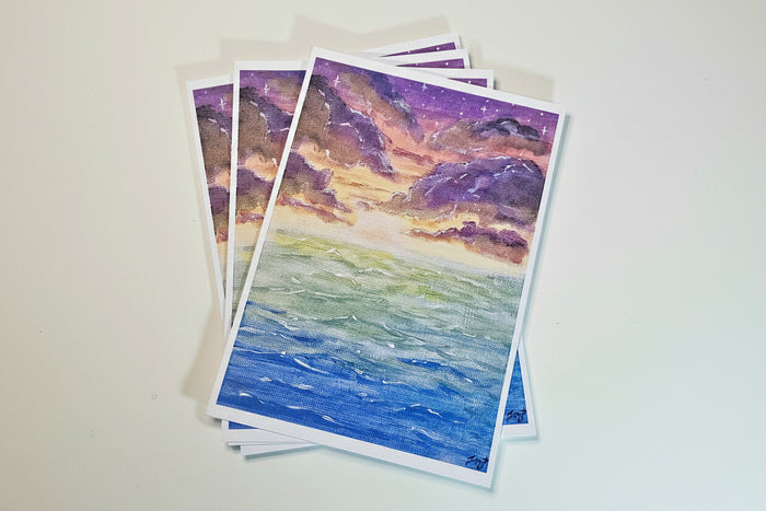 A stack of watercolor art prints depicting a coastal landscape with blue ocean waves and purple, pink, and orange mountains under a starry sky, displayed on a light beige surface.