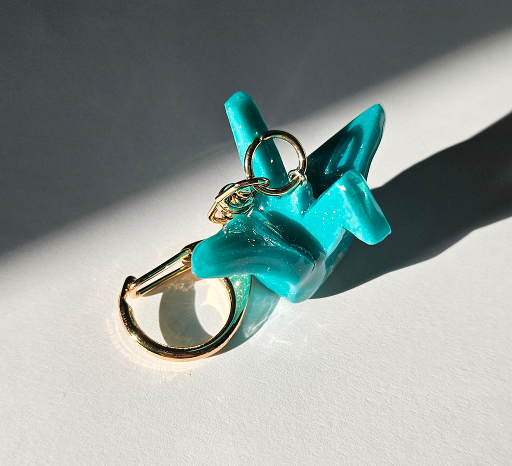Turquoise polymer clay crane keychain with a gold ring on a white background